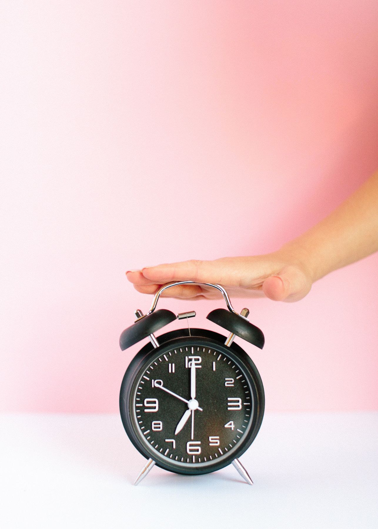 hand on top of black alarm clock with a pink background